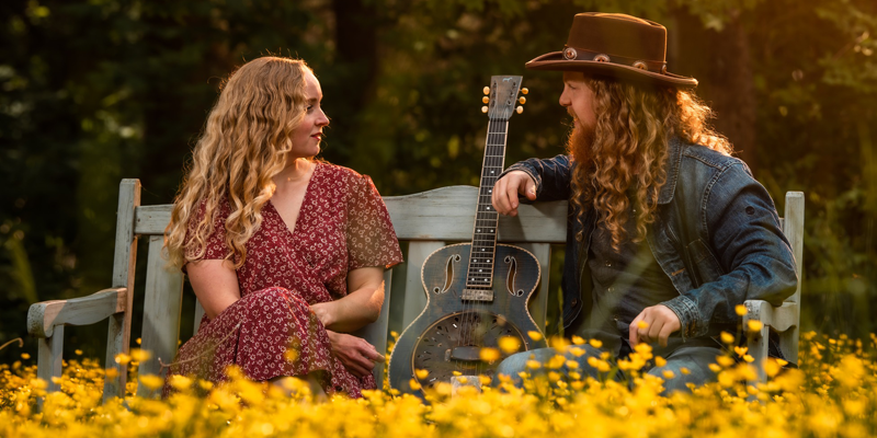 KodyandHerren Kody and Herren sitting on a bench in a field of yellow wild flowers with a guitar.